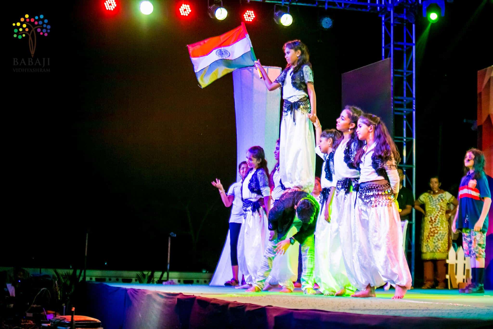 A group of girl students and a boy is performing dance with Indian flag in hand, audience are watching them behind.