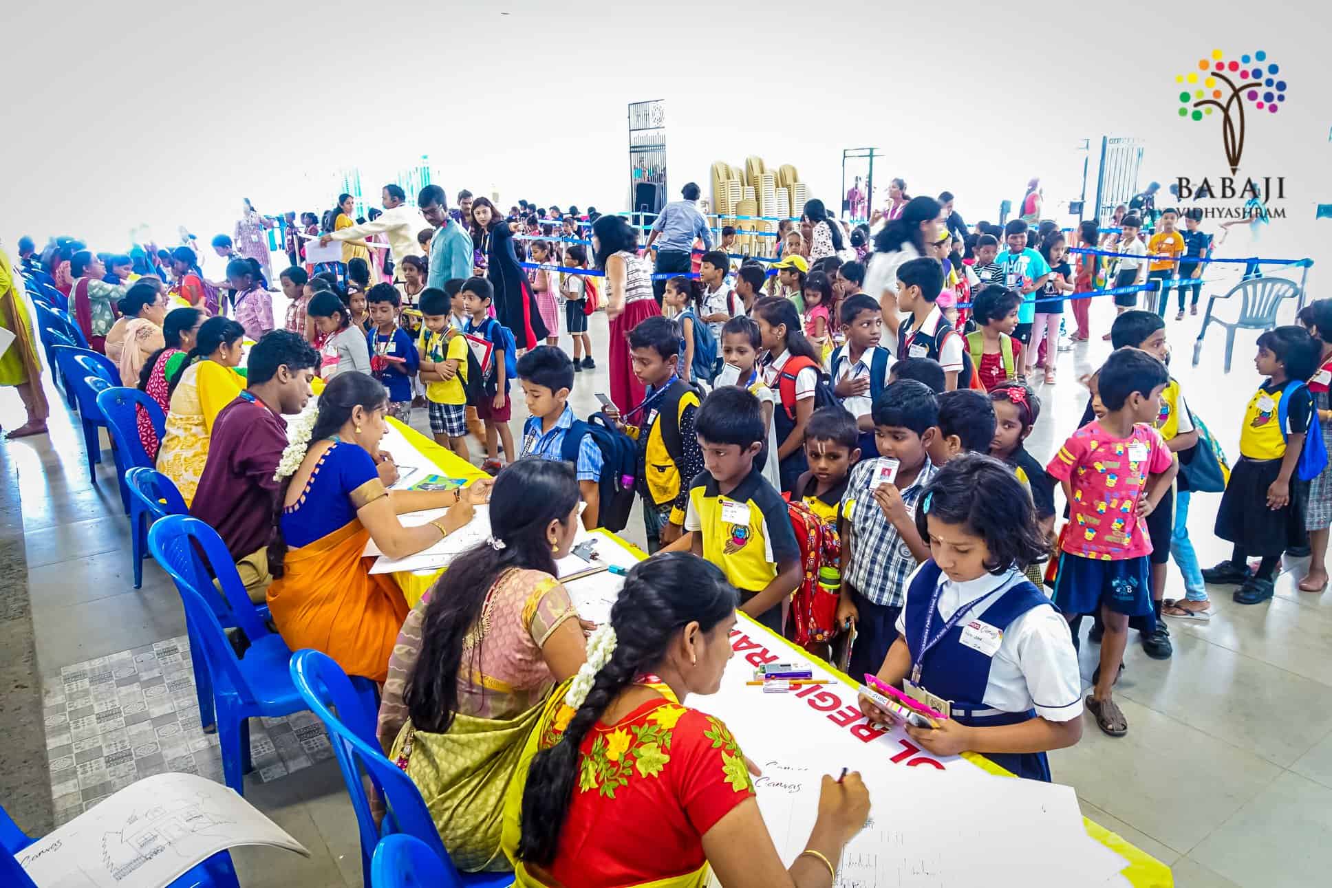 Small school kids with uniform are standing in queue and approaching teachers who are sitting in line on chairs with registration forms to enroll primary school students in cultural activities.