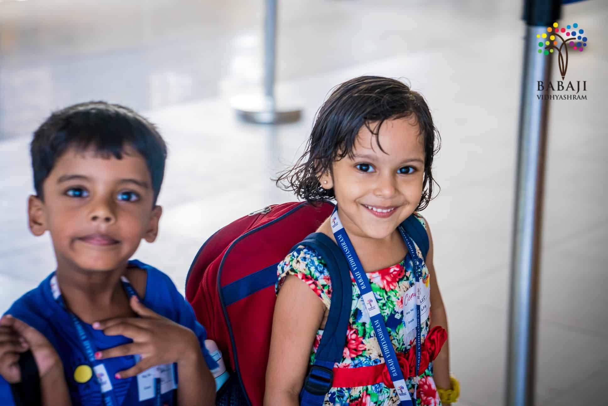Two primary section children of Babaji vidyashram with their school bags with the girl child in colourful frock and the boy child in a blue T Shirt  smiling at the camera.