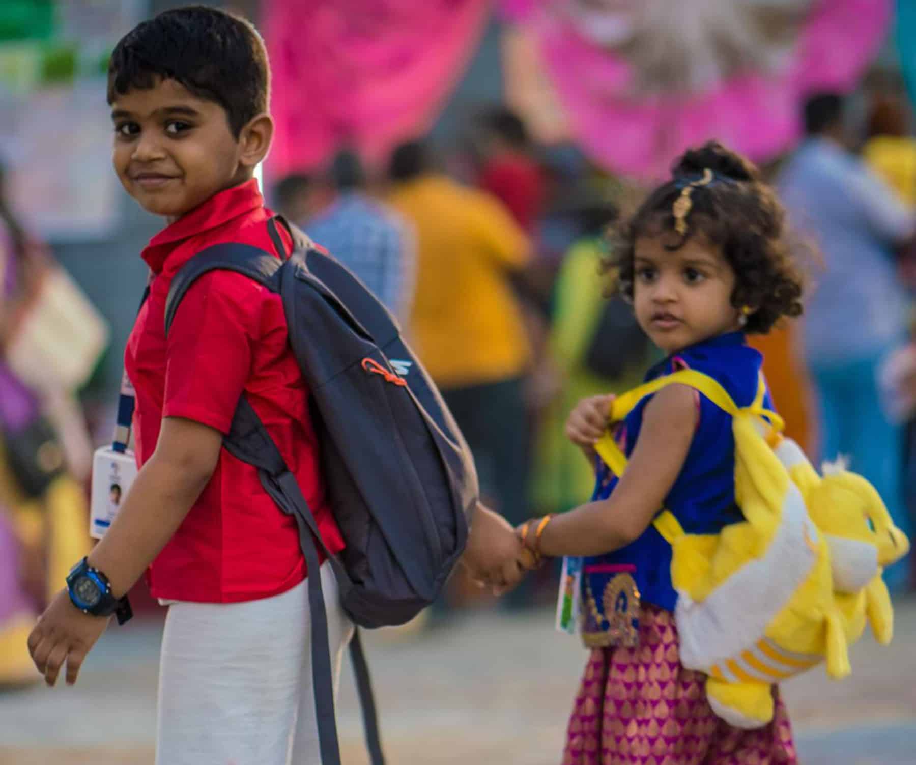 Two kids wearing traditional dresses joining their hands wearing school bags on their back on their way to school