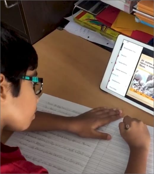 A Babaji Vidyashram School Student wearing glasses is taking down notes while attending online classes on a handheld device