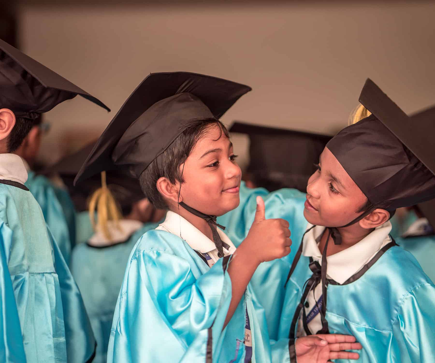 Happy Babaji Vidyashram School Children wearing graduation ceremon dress and mortarboard hat with one showing thumbs up as the other looks on.