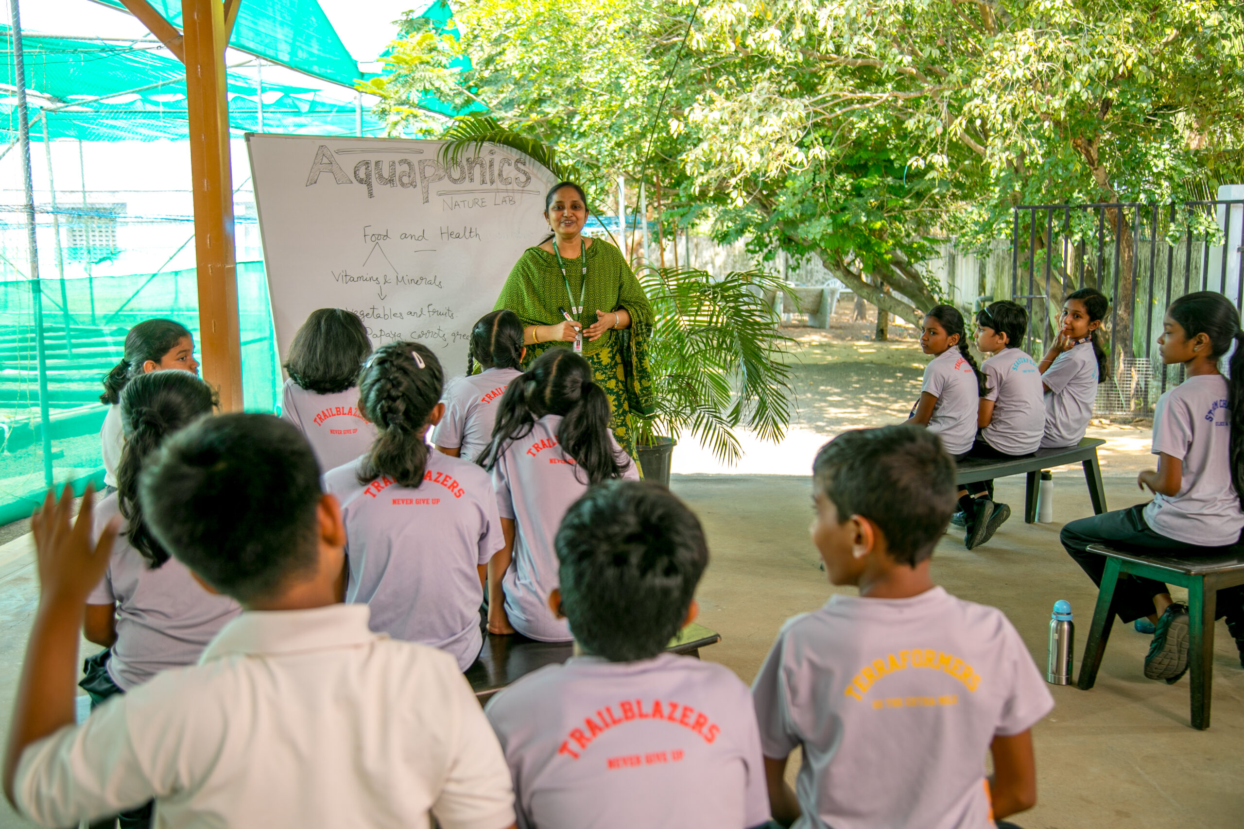 A school teacher is teaching little school students, who are seen sitting on a bench, a lesson about Aquaponics with the help of a white board and marker.